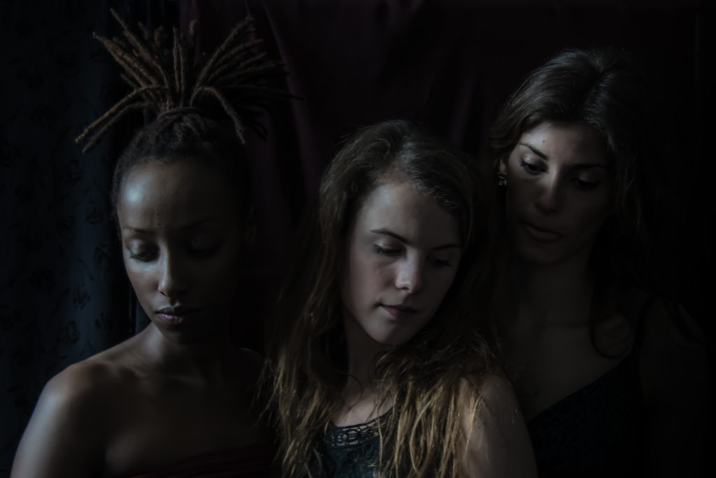Trois femmes se tiennent côte à côte dans un éclairage tamisé. La femme de gauche a des dreadlocks, celle du milieu a de longs cheveux châtain clair et la femme de droite a de longs cheveux foncés. Une photographie de Pascal Bernheim