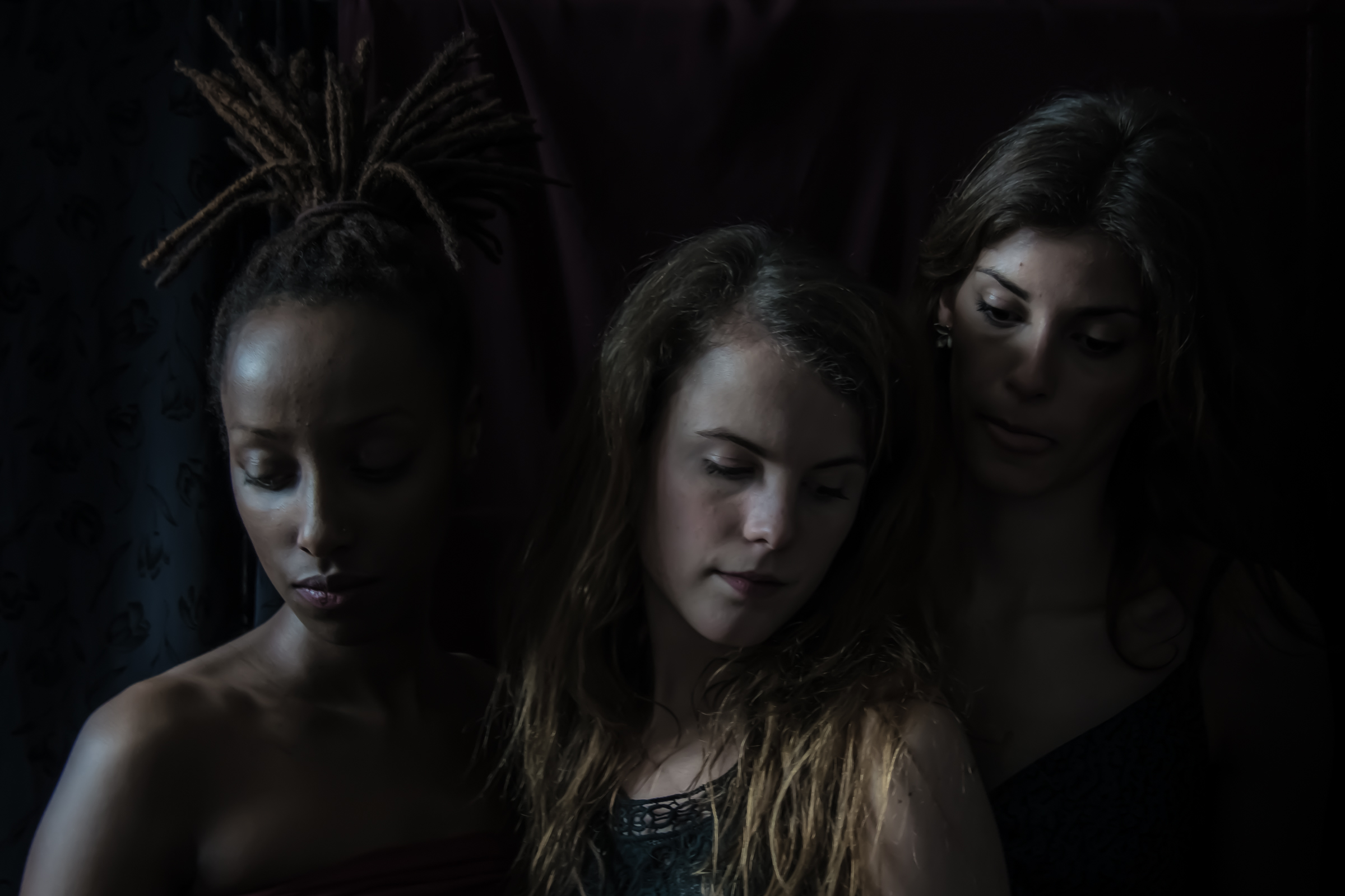 Trois femmes se tiennent côte à côte dans un éclairage tamisé. La femme de gauche a des dreadlocks, celle du milieu a de longs cheveux châtain clair et la femme de droite a de longs cheveux foncés. Une photographie de Pascal Bernheim