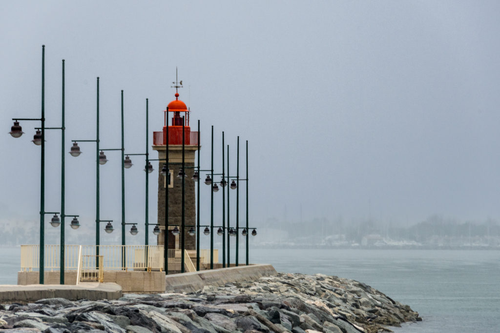 Une scène brumeuse mettant en vedette un phare au sommet rouge, entouré de poteaux métalliques avec des lumières, situé sur une jetée. Une photographie de Pascal Bernheim