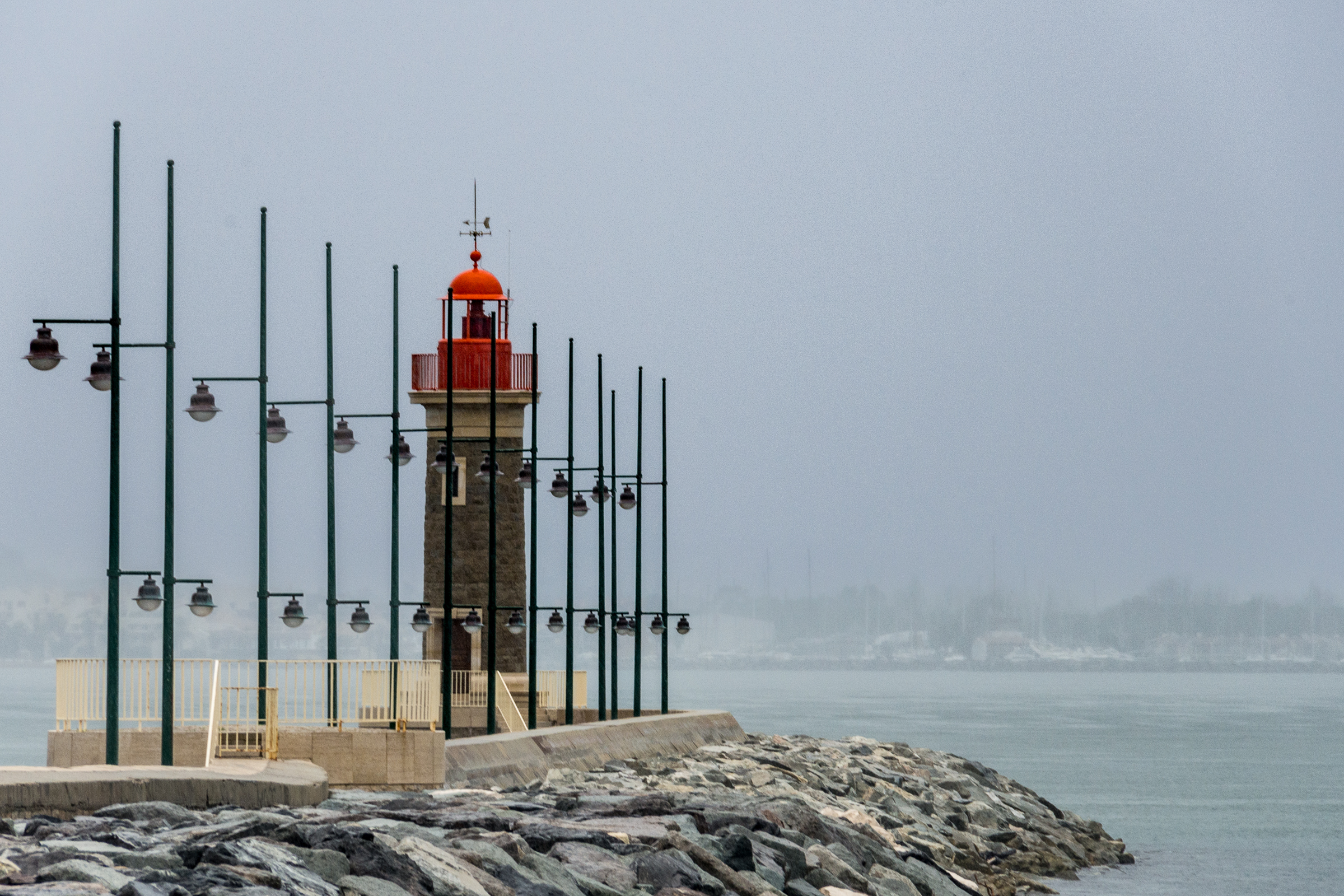 Une scène brumeuse mettant en vedette un phare au sommet rouge, entouré de poteaux métalliques avec des lumières, situé sur une jetée. Une photographie de Pascal Bernheim