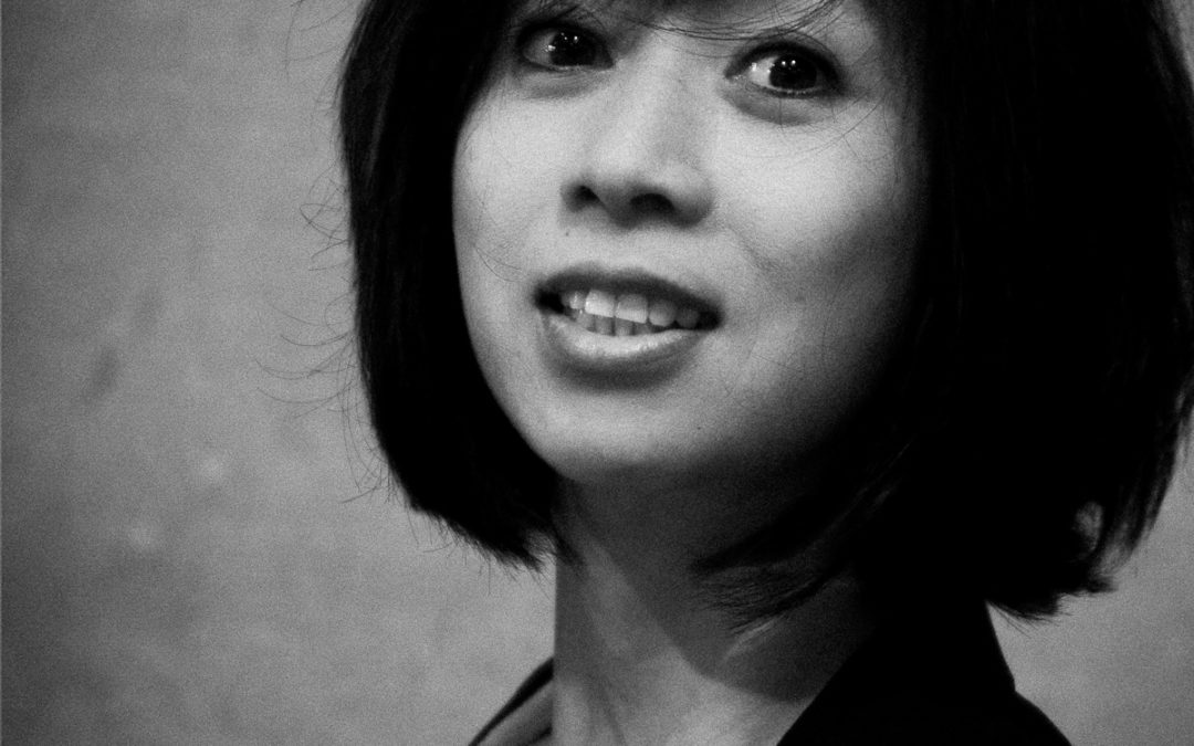 Portrait en noir et blanc de Maï, une femme aux cheveux courts et foncés, vêtue d'un blazer, regardant légèrement devant l'appareil photo avec une expression neutre. Une photographie de Pascal Bernheim