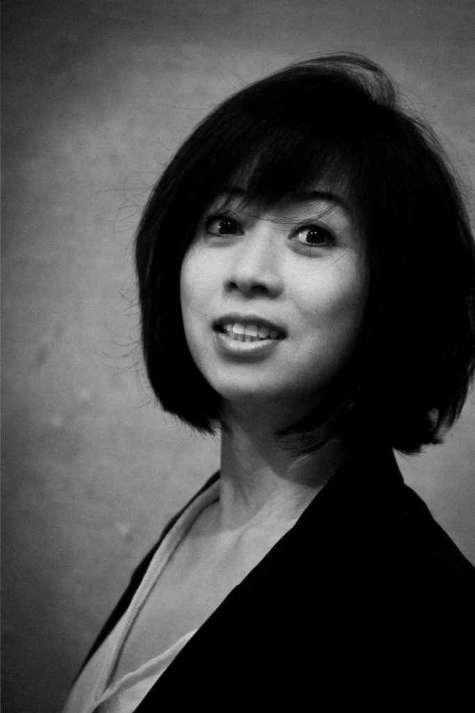 Portrait en noir et blanc de Maï, une femme aux cheveux courts et foncés, vêtue d'un blazer, regardant légèrement devant l'appareil photo avec une expression neutre. Une photographie de Pascal Bernheim