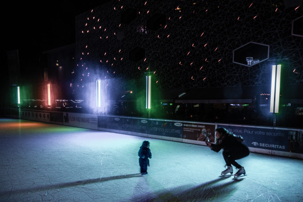 Une mère photographie son enfant sur une patinoire éclairée la nuit. L’arrière-plan présente un bâtiment éclairé avec une façade géométrique et des lumières verticales colorées. Une photographie de Pascal Bernheim