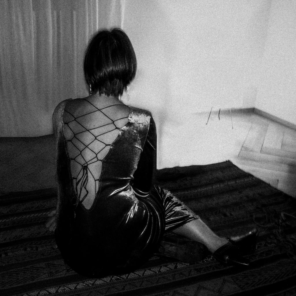 Mélanie vêtue d’une robe sombre à lacets est assise sur un tapis à motifs face à un mur dans une pièce faiblement éclairée. Une photographie de Pascal Bernheim