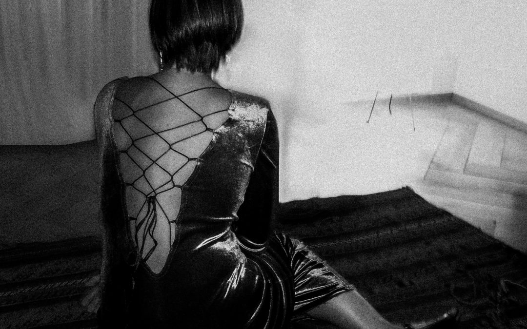 Mélanie vêtue d’une robe sombre à lacets est assise sur un tapis à motifs face à un mur dans une pièce faiblement éclairée. Une photographie de Pascal Bernheim