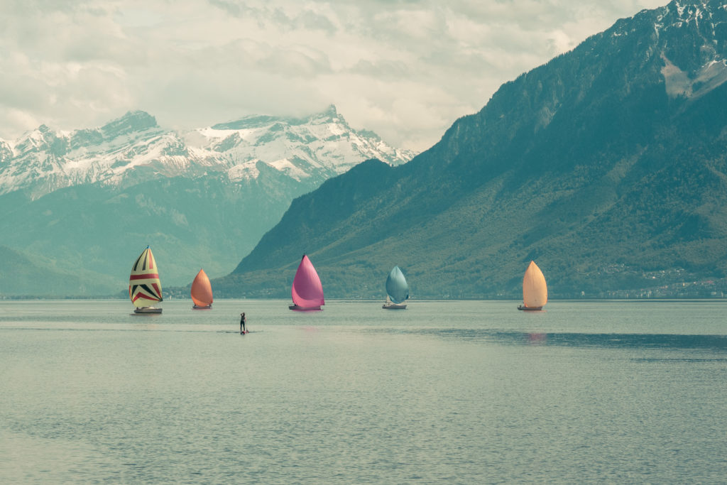 Plusieurs voiliers aux voiles colorées flottent sur le calme Lac Léman, avec des montagnes enneigées en arrière-plan et un ciel couvert. Une personne sur un paddleboard est visible au premier plan. Une photographie de Pascal Bernheim